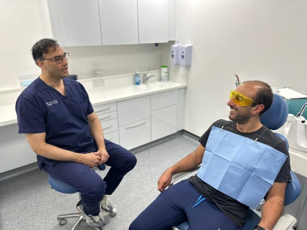 A dentist in navy scrubs is seated on a stool, having a conversation with a smiling patient wearing yellow protective glasses and a disposable dental bib in a modern dental clinic
